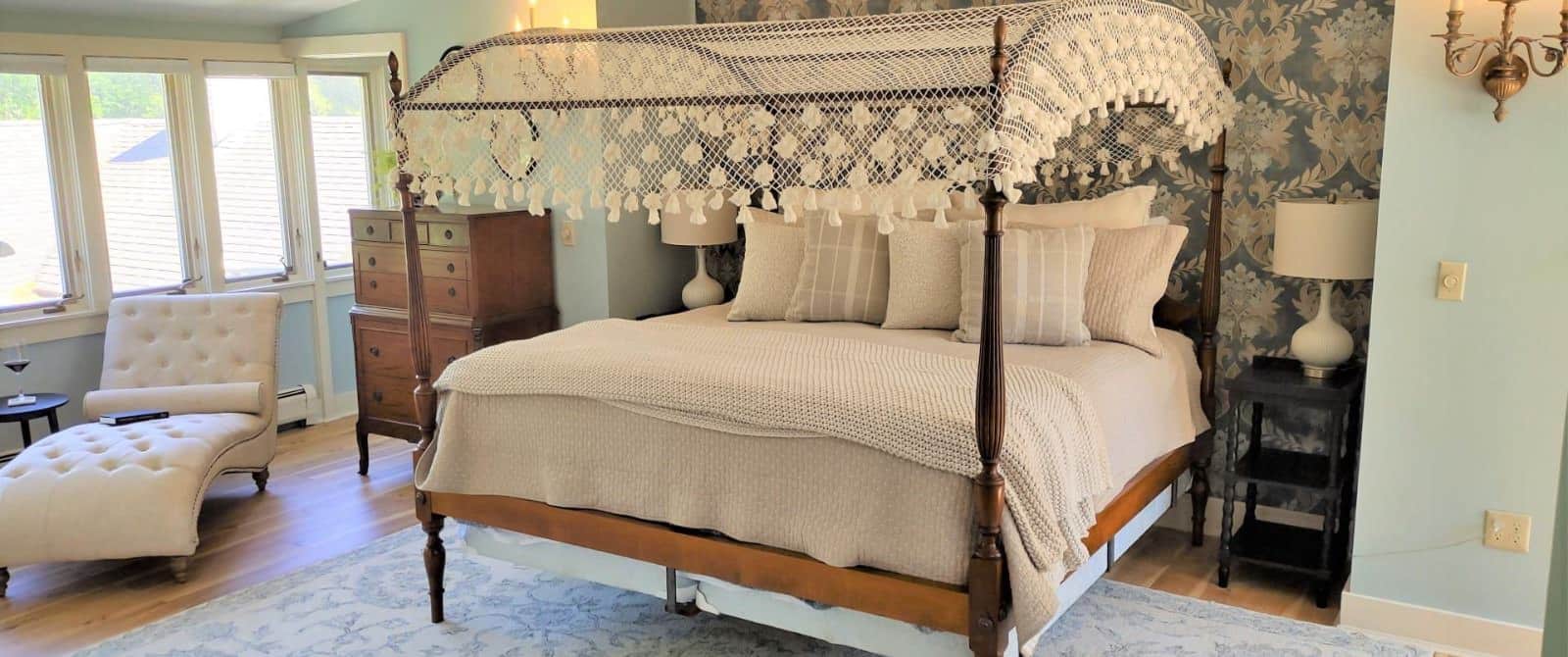 Bedroom with light blue walls, white trim, hardwood flooring, wooden four-poster bed, tan bedding, chaise, and wooden dresser