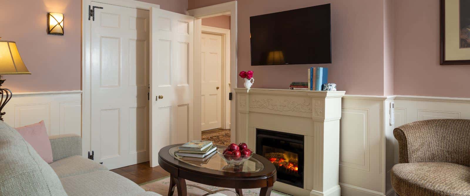 Gathering room with pink walls, white trim, upholstered sofa and chair, wooden coffee table, fireplace, and wall-mounted TV