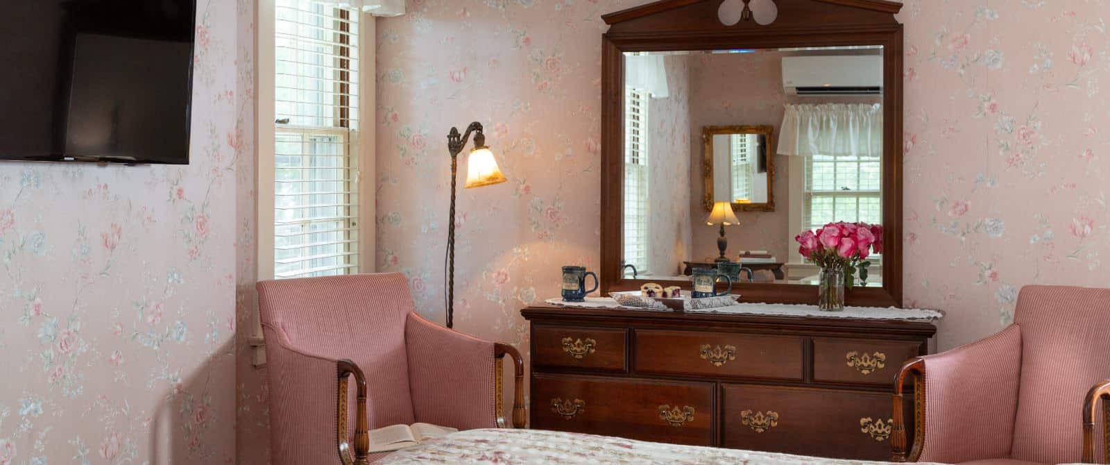 Bedroom with pink floral wall paper, floral comforter, wall-mounted flat-screen TV, dark wooden dresser with mirro, and pink upholstered antique wooden chairs