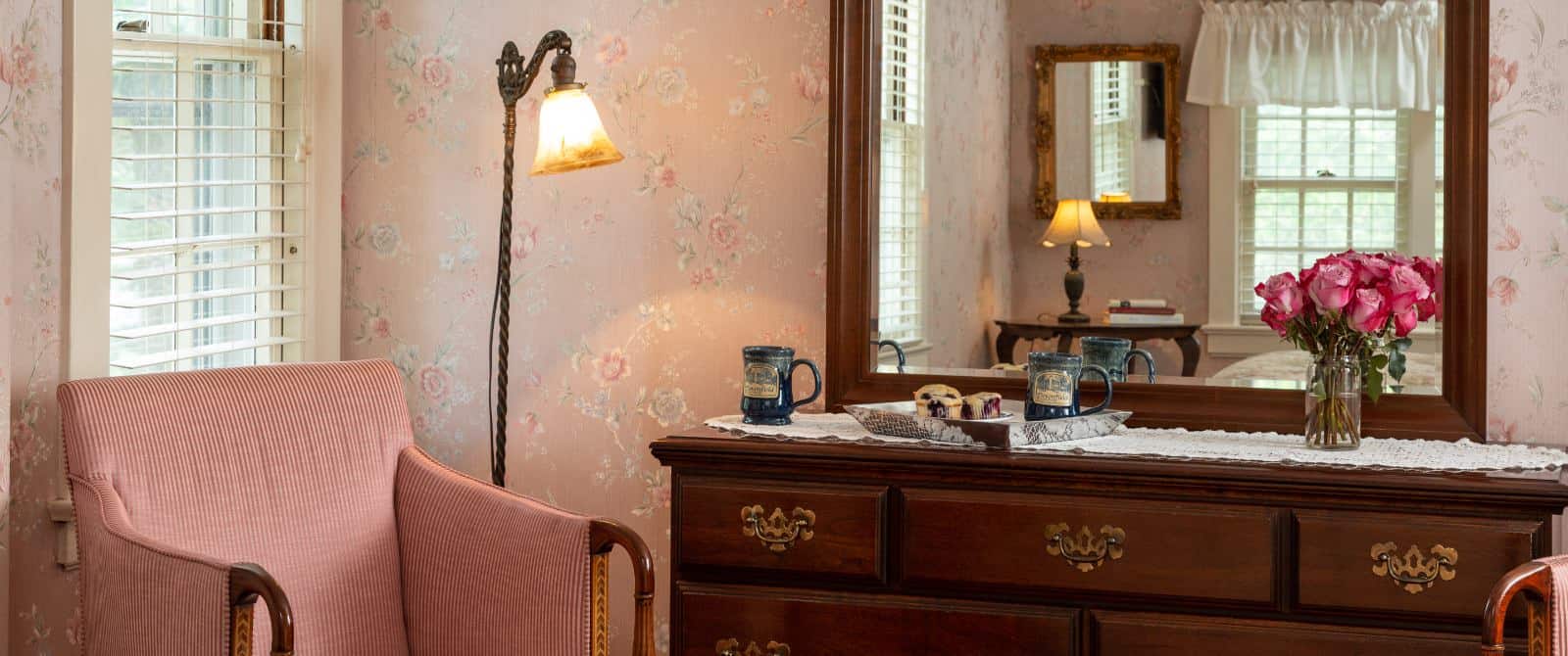 Bedroom with pink floral wall paper, dark wooden dresser with mirror, and pink upholstered antique wooden chair