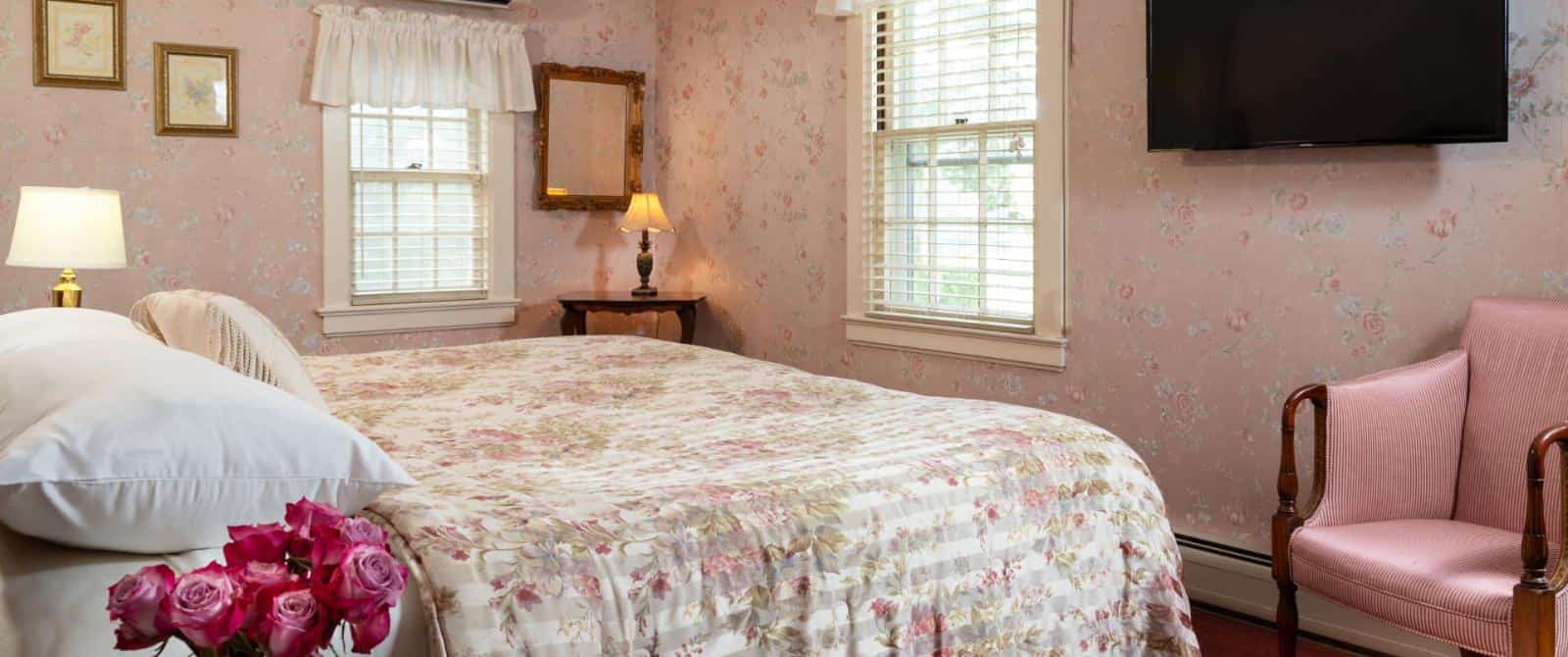 Bedroom with pink floral wall paper, floral comforter, wall-mounted flat-screen TV, and pink upholstered antique wooden chair