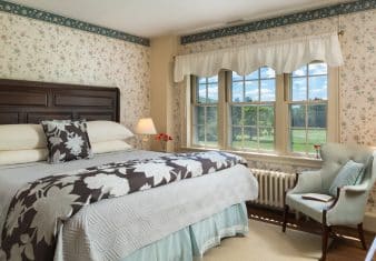 Bedroom with floral wallpaper, hardwood flooring, wooden bed, multiple hues of blue bedding, and light blue upholstered wooden chair