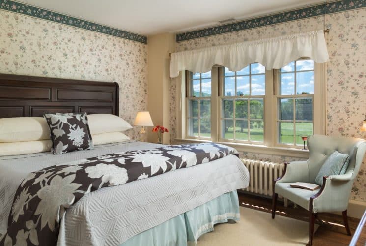 Bedroom with floral wallpaper, hardwood flooring, wooden bed, multiple hues of blue bedding, and light blue upholstered wooden chair