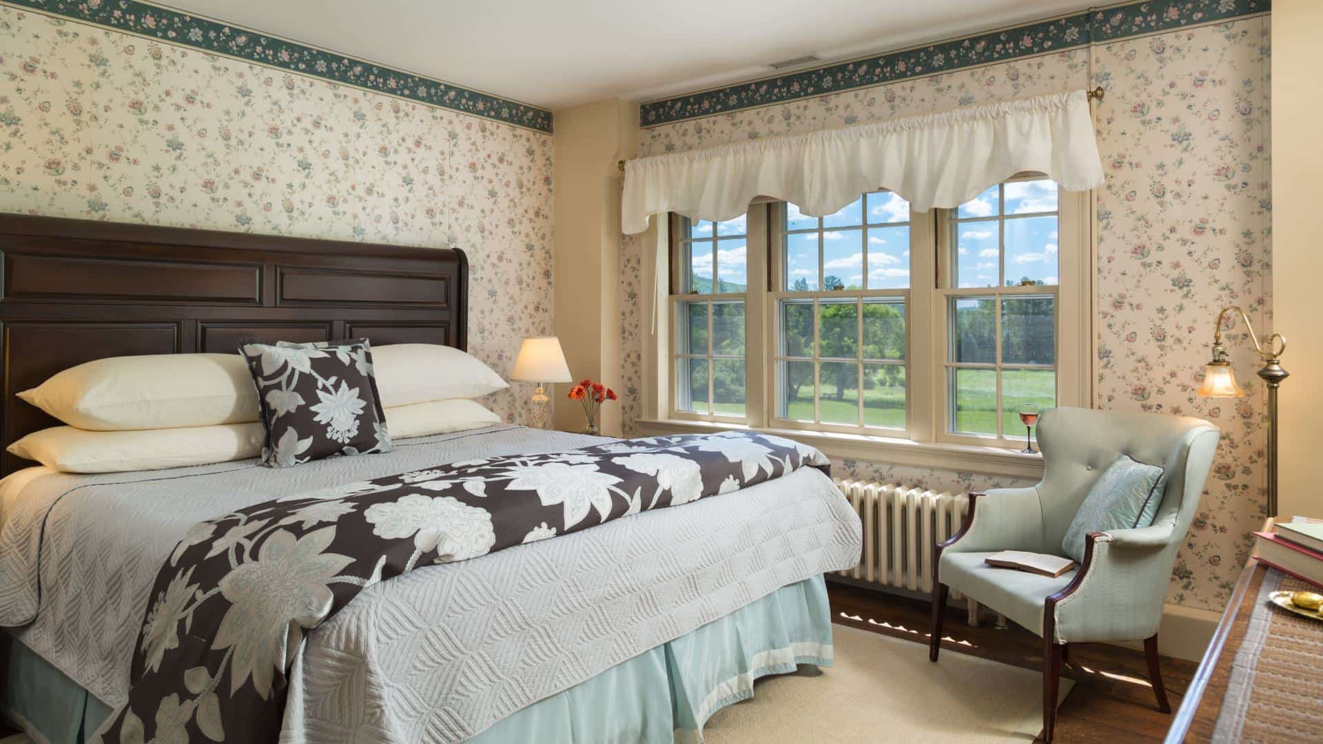 Bedroom with floral wallpaper, hardwood flooring, wooden bed, multiple hues of blue bedding, and light blue upholstered wooden chair