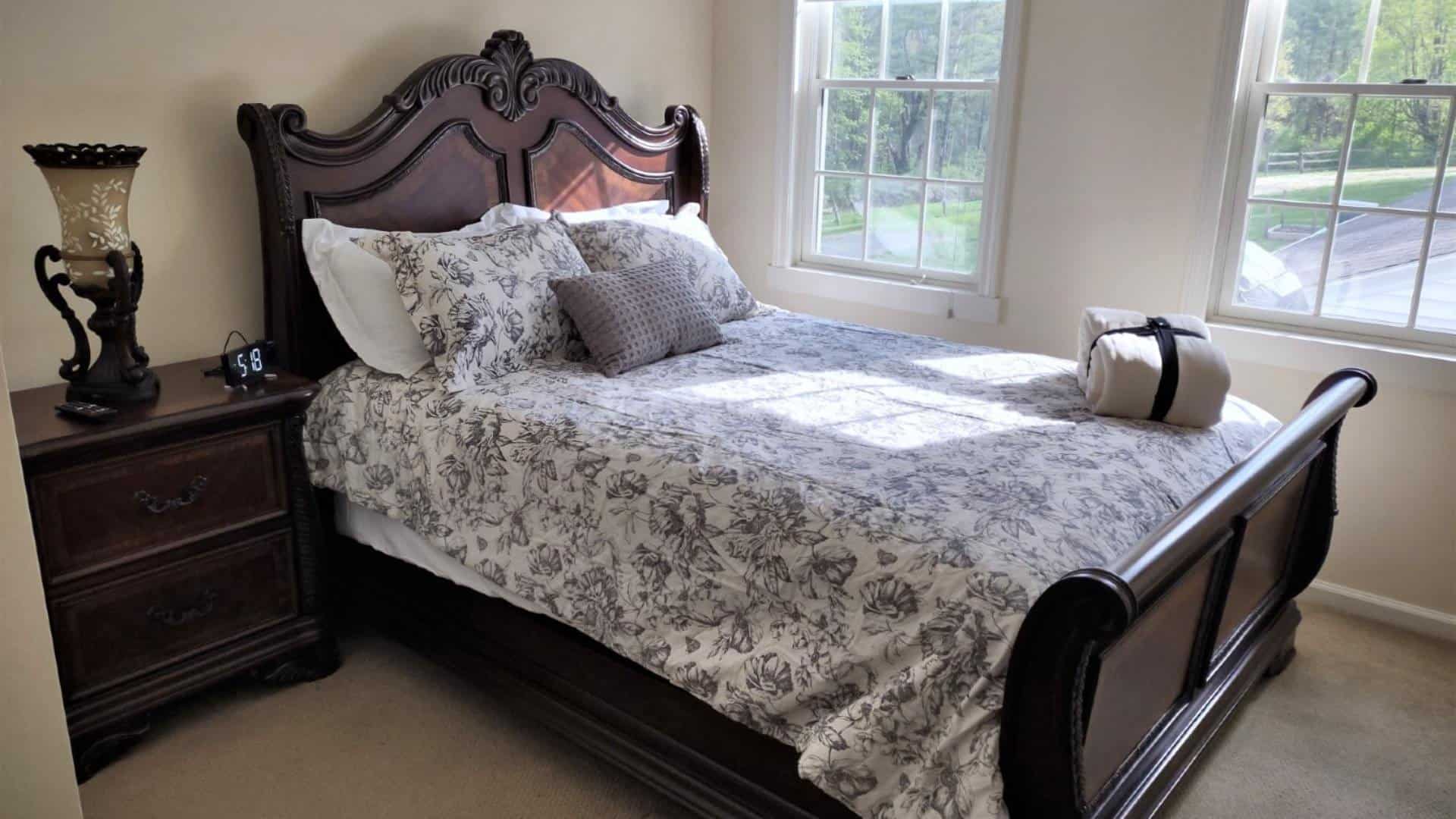 Bedroom with light colored walls and carpetting, dark wooden sleigh bed, floral bedding, wooden dresser, and windows
