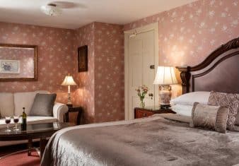 Bedroom with dark floral wallpaper, red carpeting, dark wooden headboard, gray bedding, and sitting area with upholstered sofa and dark wooden coffee table