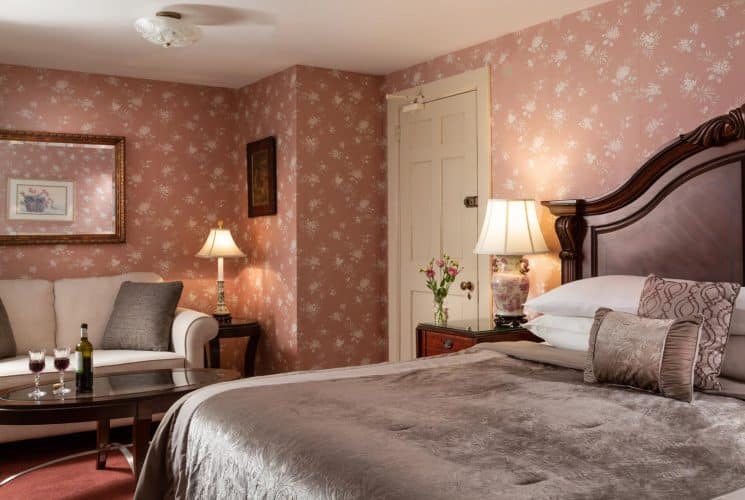 Bedroom with dark floral wallpaper, red carpeting, dark wooden headboard, gray bedding, and sitting area with upholstered sofa and dark wooden coffee table