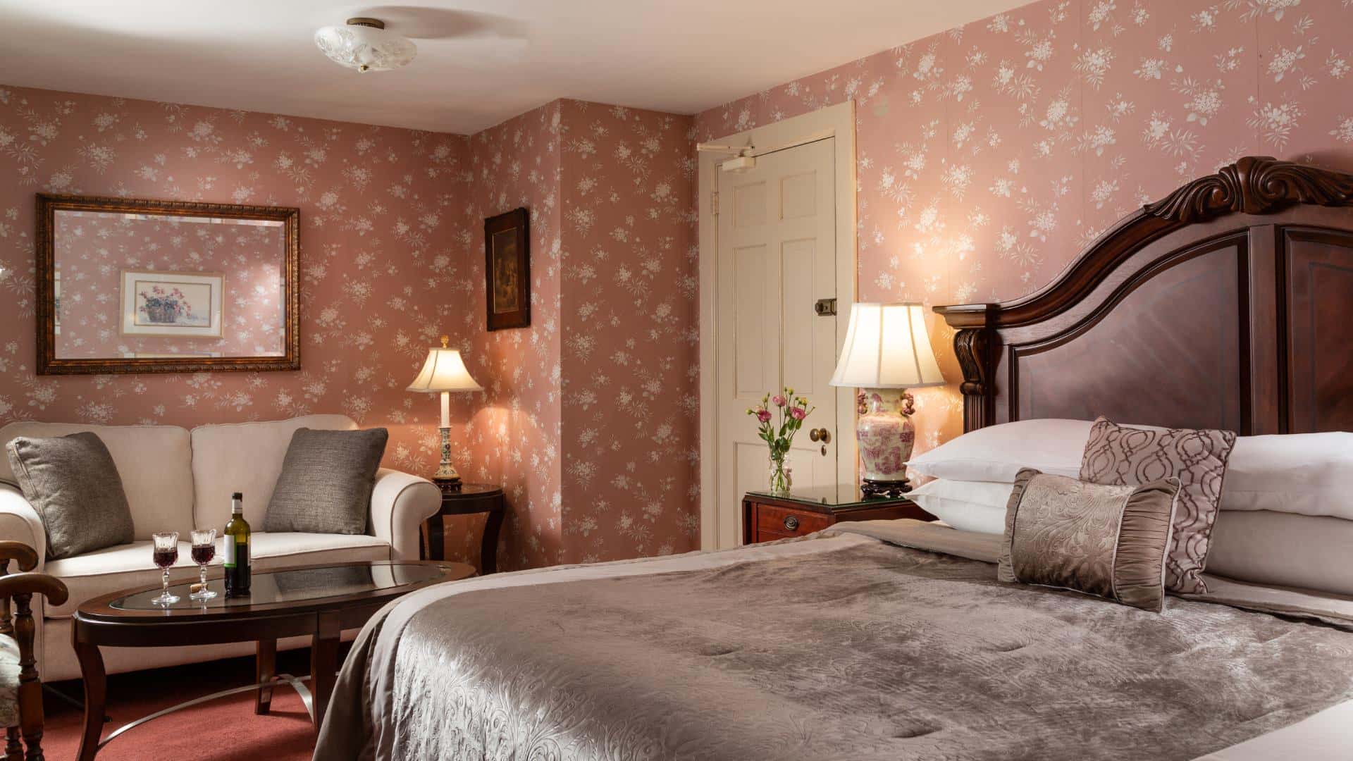 Bedroom with dark floral wallpaper, red carpeting, dark wooden headboard, gray bedding, and sitting area with upholstered sofa and dark wooden coffee table