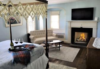 Bedroom with blue walls, white trim, hardwood flooring, dark wooden four-poster bed, lavender bedding, fireplace, wall-mounted TV, and sitting area with upholstered loveseat and wooden coffee table