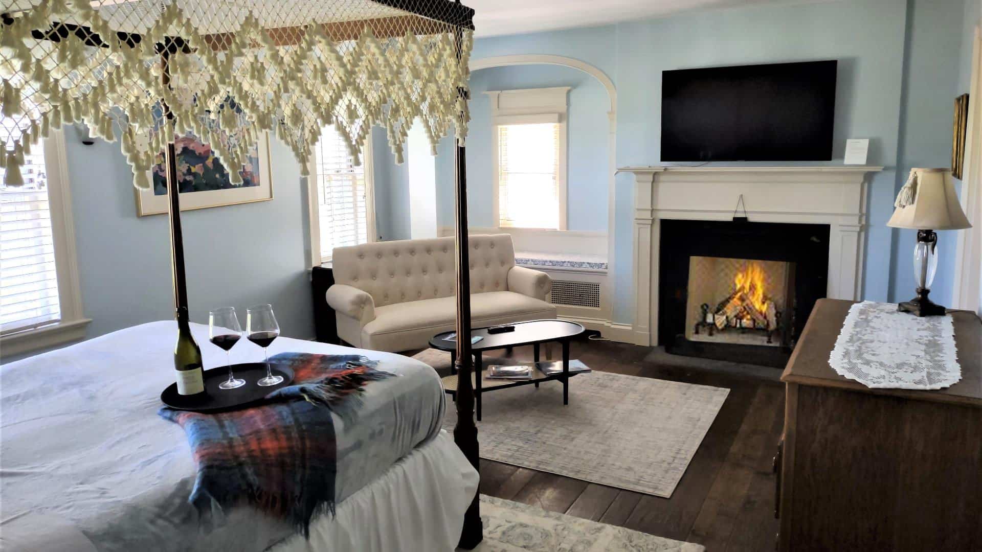 Bedroom with blue walls, white trim, hardwood flooring, dark wooden four-poster bed, lavender bedding, fireplace, wall-mounted TV, and sitting area with upholstered loveseat and wooden coffee table