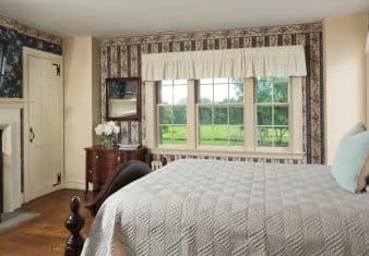 Bedroom with floral and striped wallpaper, hardwood flooring, wooden bed, light gray bedding, fireplace, and large window