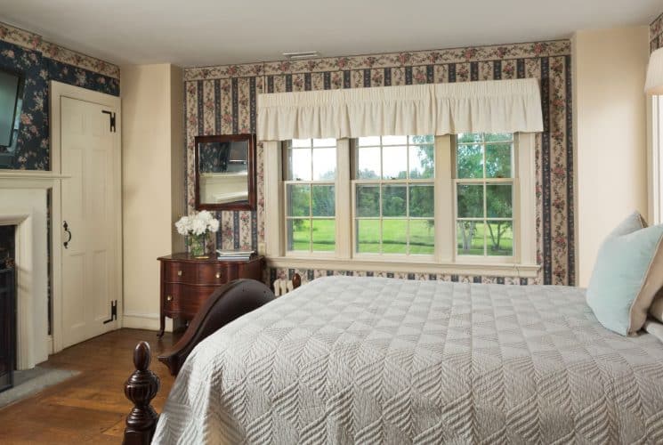 Bedroom with floral and striped wallpaper, hardwood flooring, wooden bed, light gray bedding, fireplace, and large window