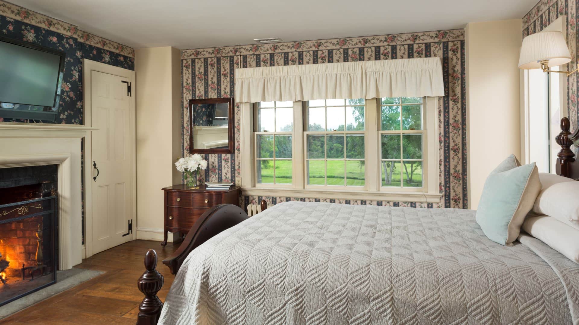 Bedroom with floral and striped wallpaper, hardwood flooring, wooden bed, light gray bedding, fireplace, and large window