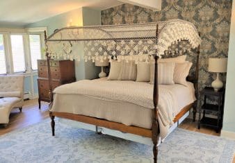 Bedroom with light blue walls, white trim, hardwood flooring, wooden four-poster bed, tan bedding, chaise, and wooden dresser