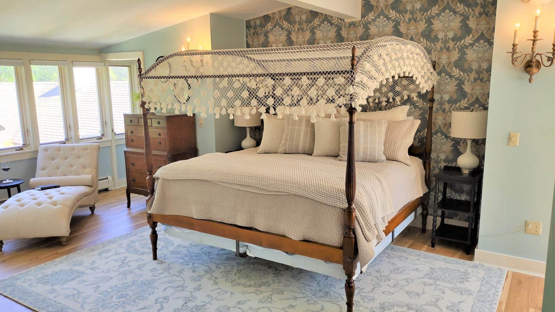 Bedroom with light blue walls, white trim, hardwood flooring, wooden four-poster bed, tan bedding, chaise, and wooden dresser