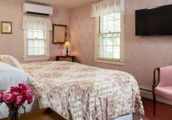 Bedroom with pink floral wall paper, floral comforter, wall-mounted flat-screen TV, and pink upholstered antique wooden chair