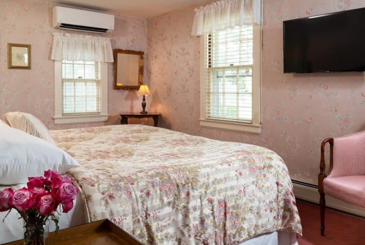 Bedroom with pink floral wall paper, floral comforter, wall-mounted flat-screen TV, and pink upholstered antique wooden chair
