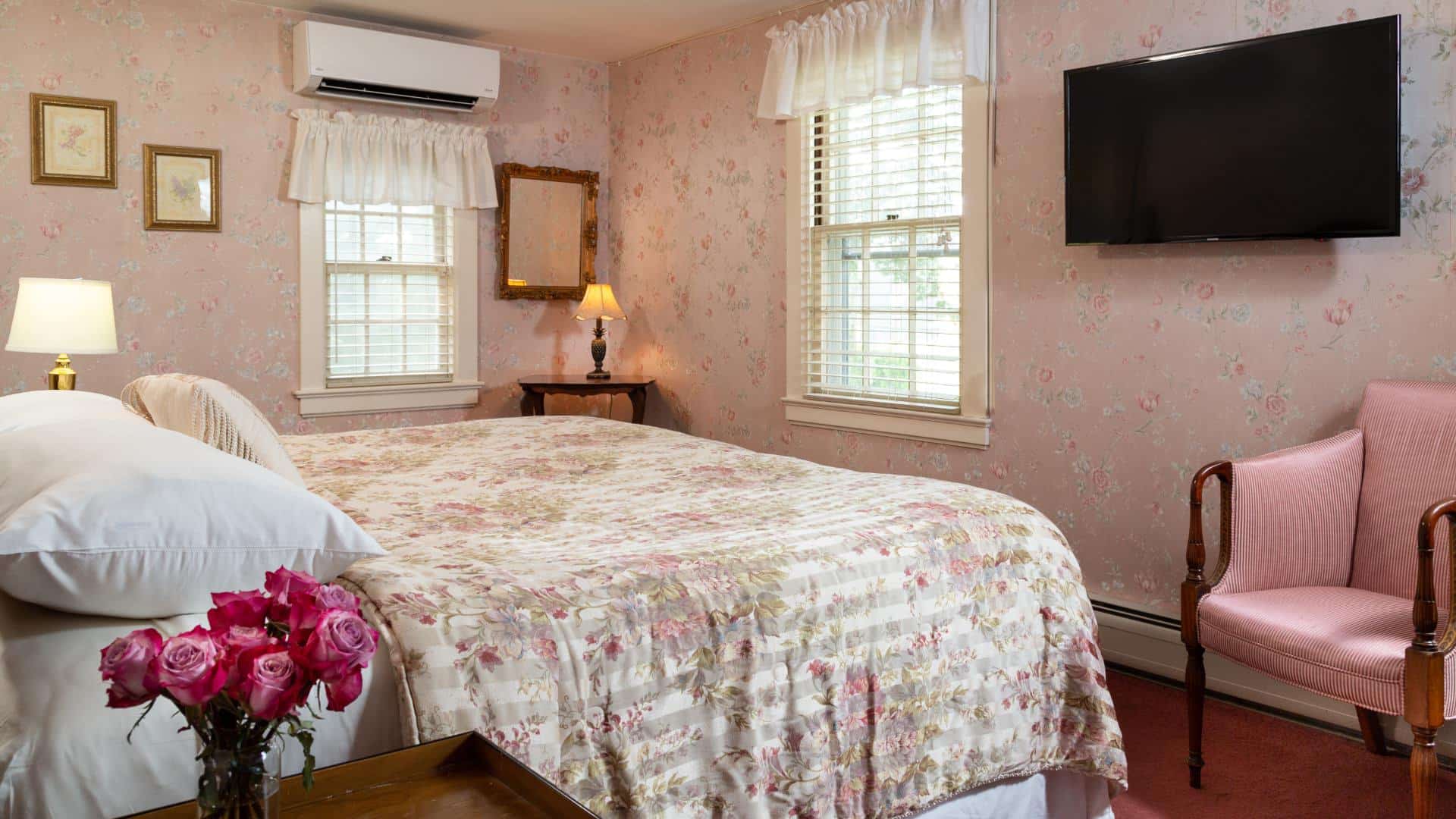 Bedroom with pink floral wall paper, floral comforter, wall-mounted flat-screen TV, and pink upholstered antique wooden chair