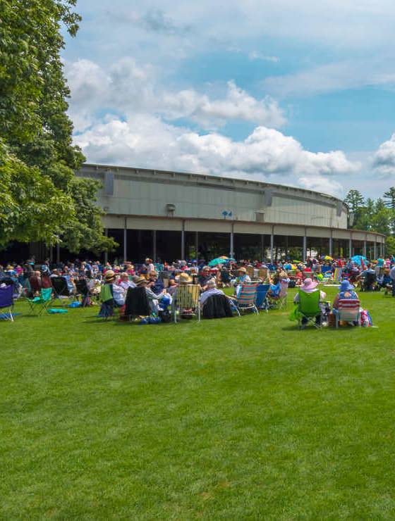 Large building surrounded by green grass and large trees with people sitting on blankets enjoying a music festival