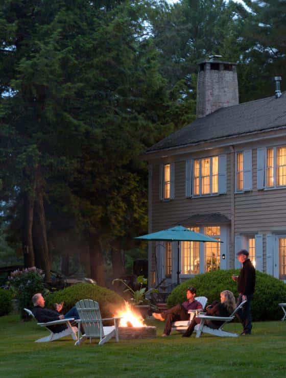 Exterior view of the property at dusk surrounded by green trees, shrubs and grass with people sitting around a firepit