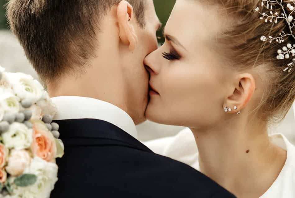 Woman in white dress holding bouquet kissing man with black suit on the cheek