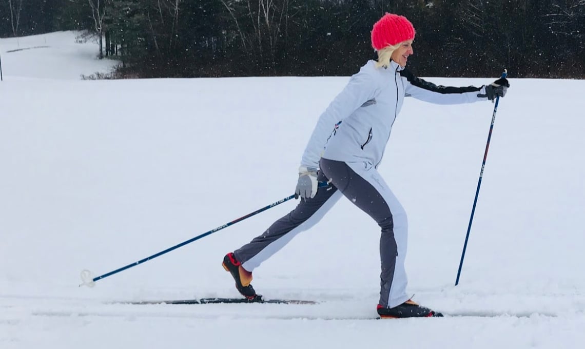 Woman in white snow suit and pink hat cross-country skiing with trees in the background