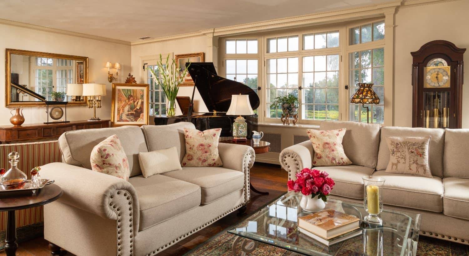 Large gathering room with cream walls, hardwood flooring, upholstered sofas, glass coffee table, grand piano, and grandfather clock