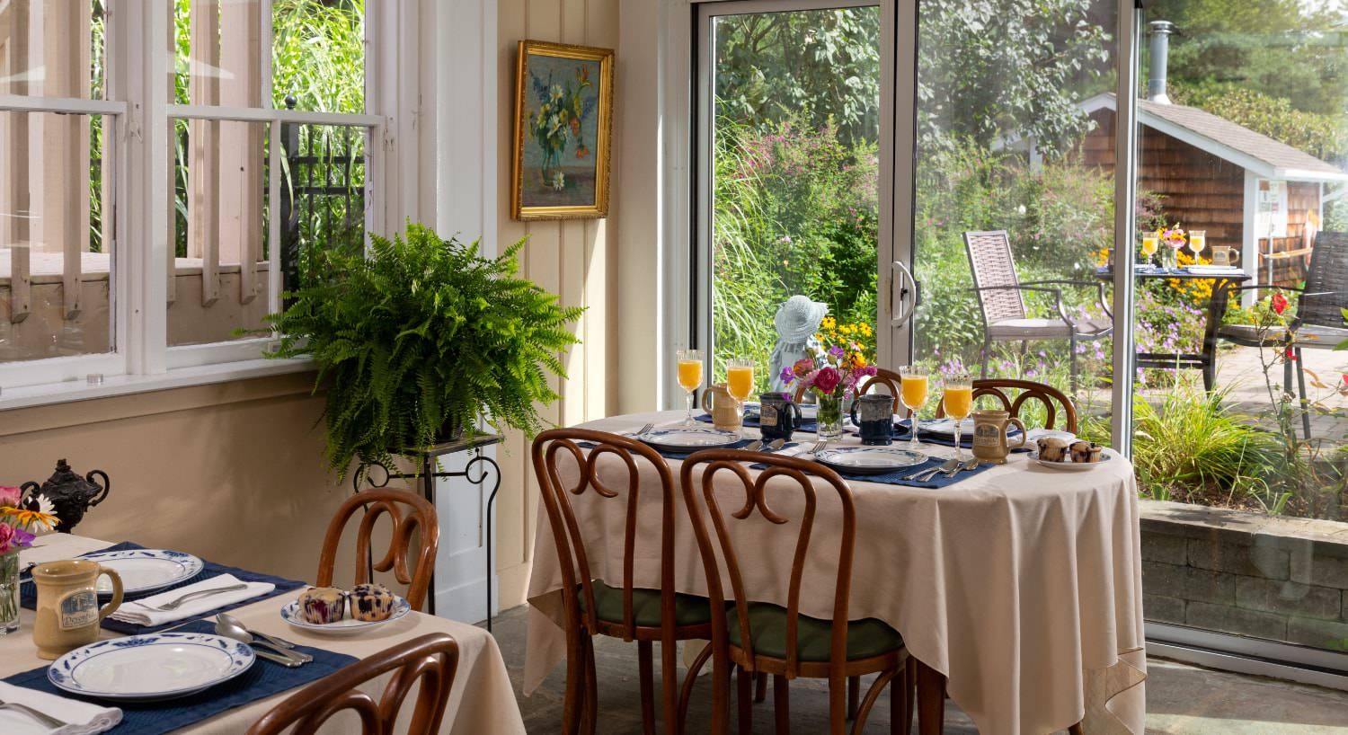 Tables with cream tablecloth set up for breakfast in a room with large sliding glass door and view to the patio surrounded by green vegetation
