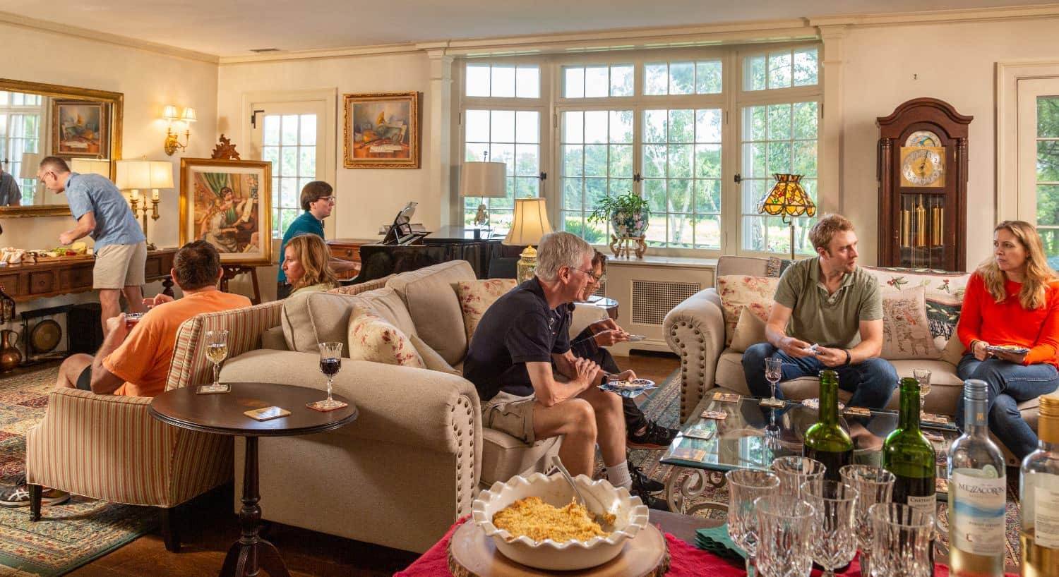 Large gathering room with cream walls, hardwood flooring, upholstered sofas, glass coffee table, grand piano, grandfather clock, and people socializing