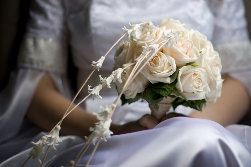 Bride in white dress sitting while holding a bouquet of cream roses